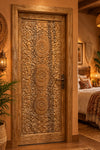 A large, intricately carved wooden door panel leaning against other carved wooden doors. The main panel features three large sunburst medallions surrounded by detailed floral and vine carvings,  